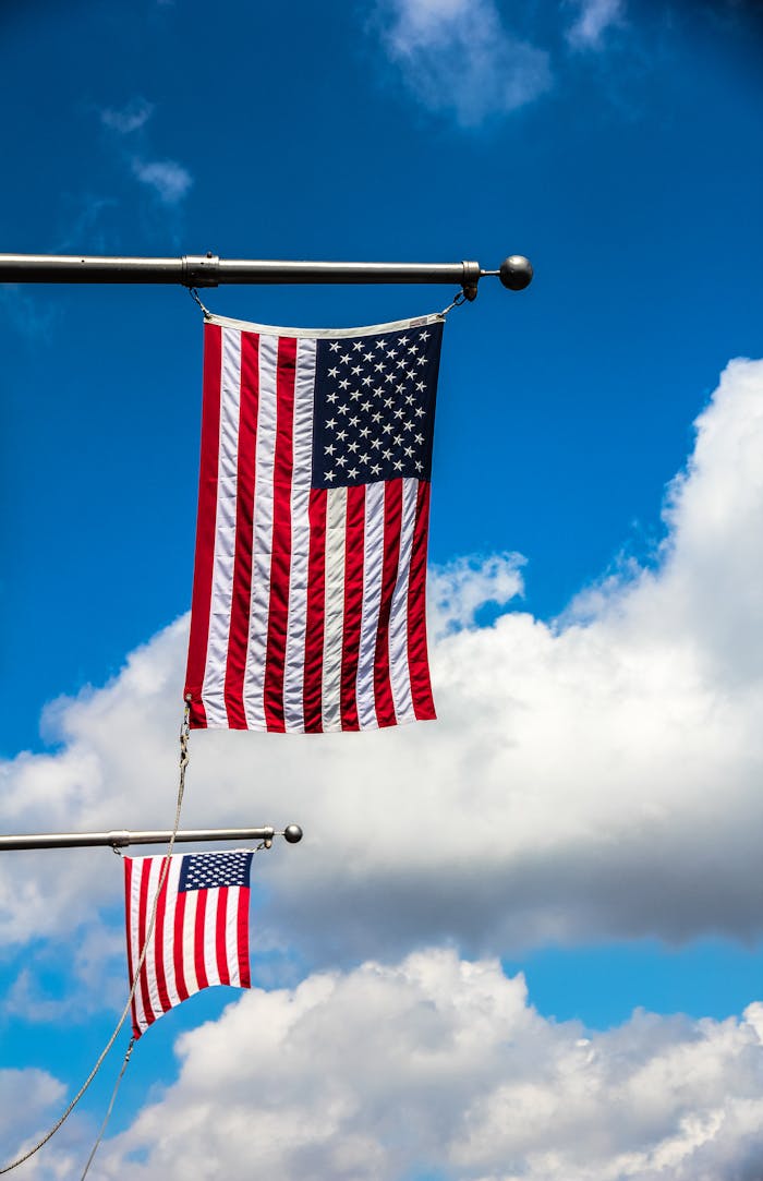 Two American flags on poles with a clear blue sky and clouds backdrop, symbolizing patriotism.